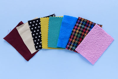 Seven fabric swatches for jackets including wool coating, tweed and twill are arranged in a fan shape on a light blue background. The swatches include solid burgundy, solid beige, black with white polka dots, mustard yellow, teal, bright blue, and a multicolor houndstooth pattern, ending with a solid pink swatch.