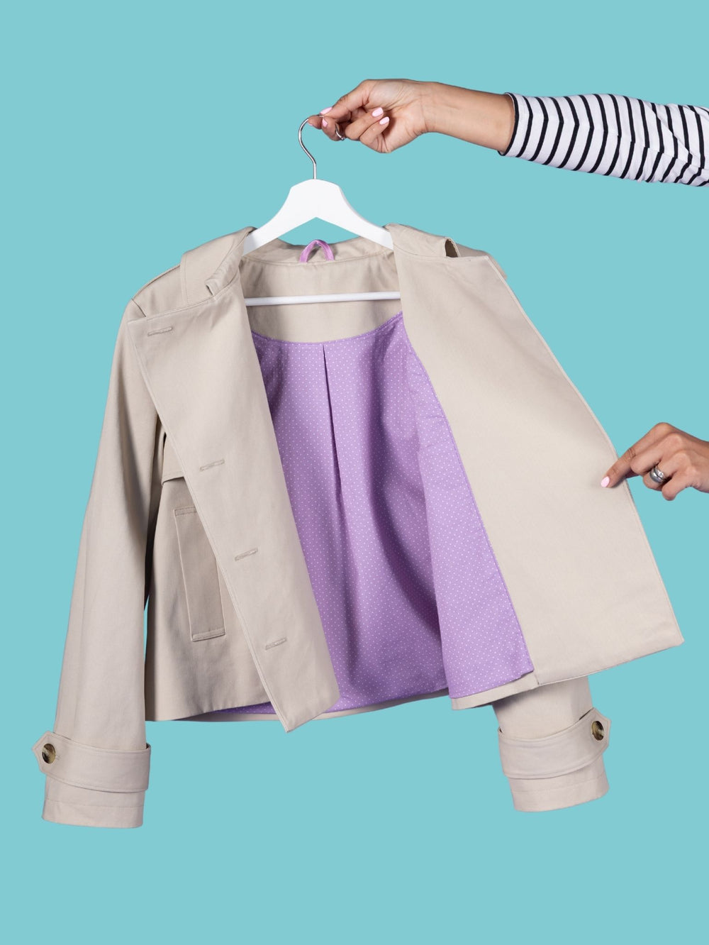 A beige Manon trench jacket is shown on a white hanger against a solid turquoise background, with a purple lining. A person's hands and striped sleeve arm are visible holding the hanger and opening the jacket.
