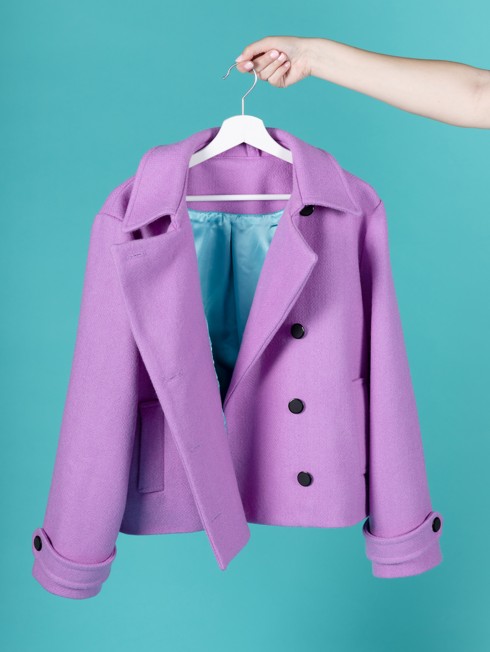 A purple double-breasted Manon pea-coat with black buttons, hanging on a white hanger held by a person's hand against a solid teal background.